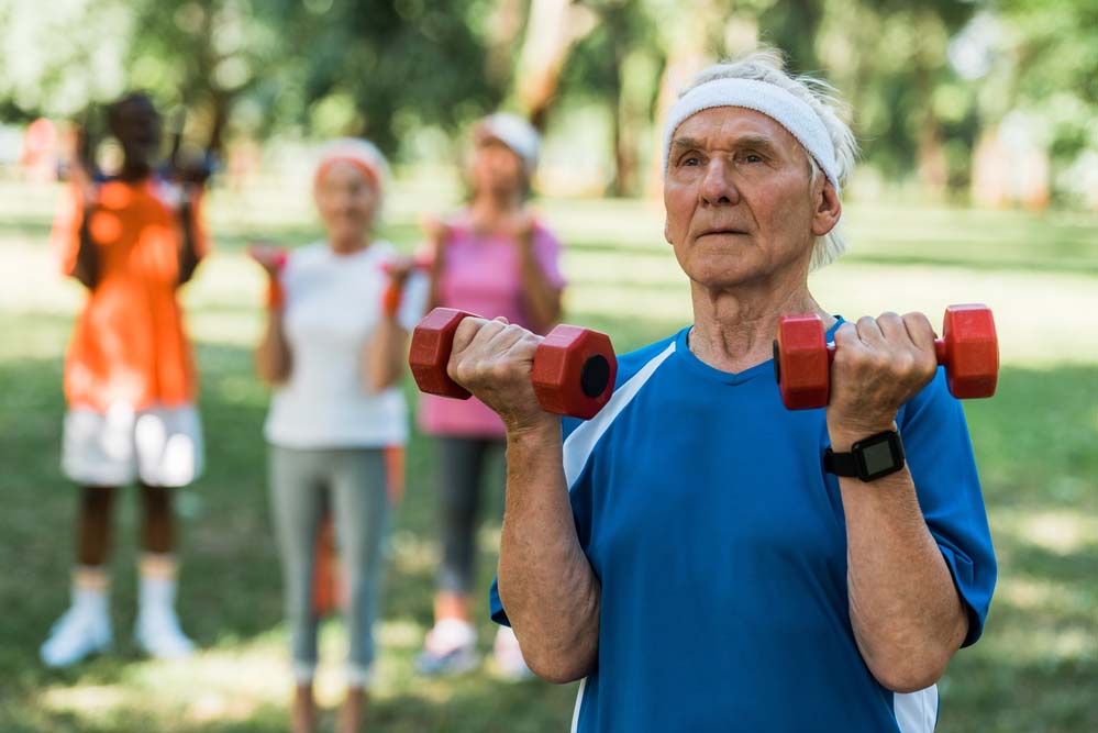 A man with a sweatband lifts two red dumbbells outside, with other people behind him and out of focus.
